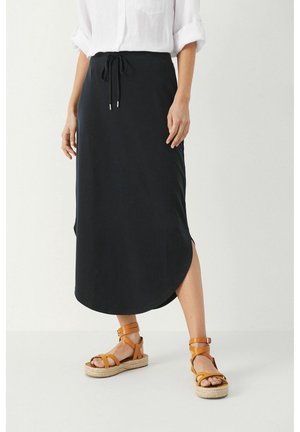 Kobieta w czarnej spódnicy midi z bocznymi rozcięciami, białej koszuli z podwiniętymi rękawami oraz beżowych, platformowych sandałach na paskach, stojąca na białej podłodze.