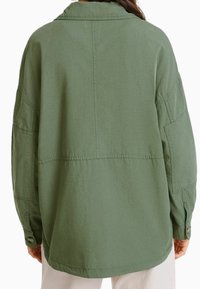 Osoba ubrana w luźną, oliwkowozieloną kurtkę z długim rękawem widzianą od tyłu, w połączeniu z jasnymi spodniami.