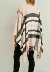 Femme portant un poncho beige avec des motifs à carreaux noirs, blancs et roses, avec des bords à franges, associé à un pantalon noir, de dos.