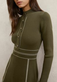 Olivzaļas krāsas ribota kleita ar augstu apkakli, baltiem šuvju akcentiem un četriem apaļiem pogām priekšpusē. Garas piedurknes.