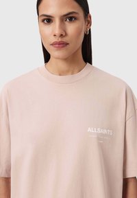 Lichtroze oversized katoenen T-shirt met een ronde hals, korte mouwen en witte logo-details aan de voorkant. Gladde textuur.