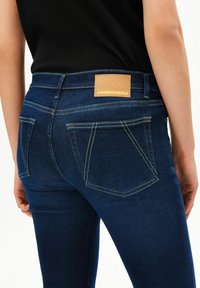 Rückansicht einer Person, die dunkelblaue Jeans mit beiger Naht und Lederetikett der Marke "ARMEDANGELS" am Bund sowie ein schwarzes Oberteil trägt.