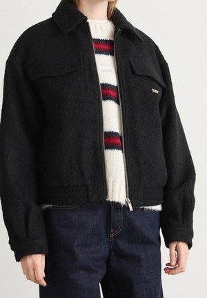 Person iført en sort uldjakke med lynlås over en hvid sweater med røde og marineblå striber, kombineret med mørkeblå denimjeans.