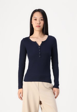 Langärmliges marineblaues Oberteil mit einem knöpfbaren Henley-Ausschnitt, das eine glatte Textur und eine figurbetonte Passform aufweist, kombiniert mit hellbeigen Hosen.