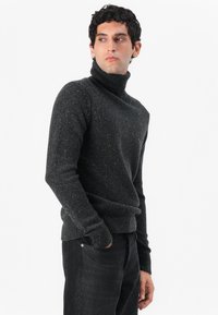 Oglata turtleneck pulover iz ogljene barve z teksturirano površino, z rebrastimi manšetami in robom, v kombinaciji s temnimi kavbojkami. Diskretni pikčasti vzorec po celotni površini.