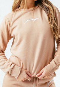 Světle béžová mikina s dlouhými rukávy, žebrovanými manžetami a kulatým výstřihem. Na přední straně se nachází bílé vyšívané text. Měkká, strukturovaná látka.