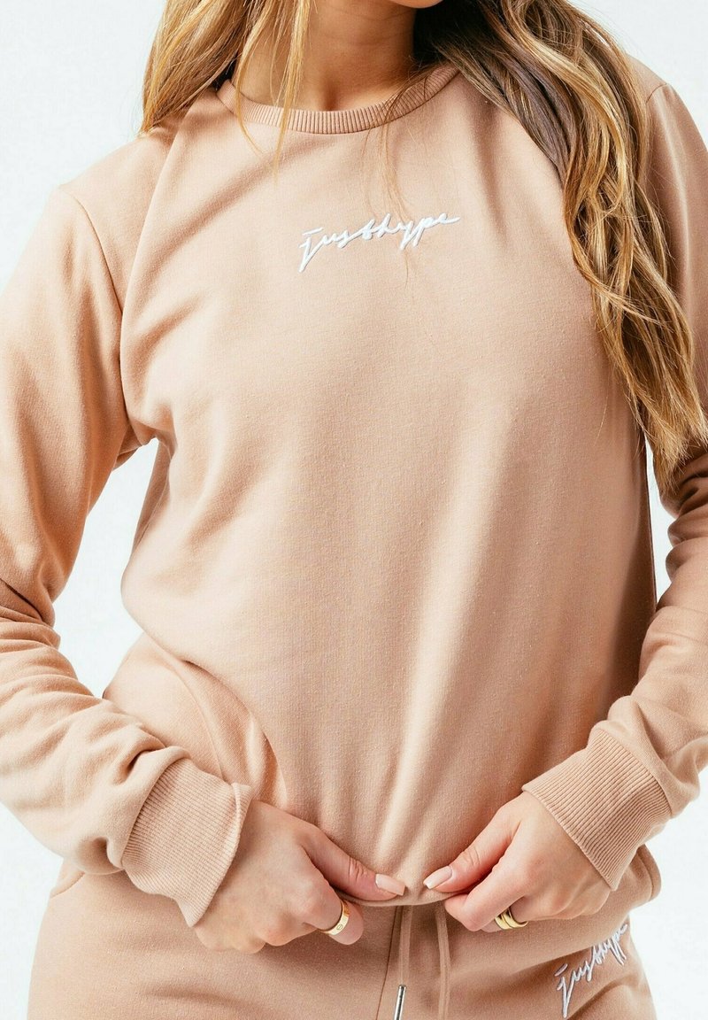 Světle béžová mikina s dlouhými rukávy, žebrovanými manžetami a kulatým výstřihem. Na přední straně se nachází bílé vyšívané text. Měkká, strukturovaná látka.