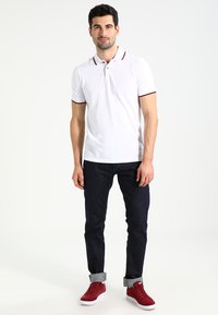 Polo bianco con colletto, accentuato da rifiniture bordeaux, abbinato a jeans blu scuro e sneakers rosse. Design semplice e aderente.