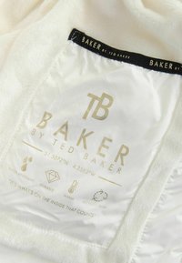 Etikette indeni på hvidt stof viser "Baker by Ted Baker" branding, koordinater og ikoner for vandmodstand, holdbarhed, varme og genanvendt fyld.