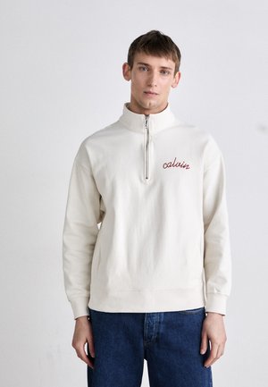 Cream-colored half-zip pullover with embroidered red "calvin" logo, made from soft material. Paired with blue denim jeans.