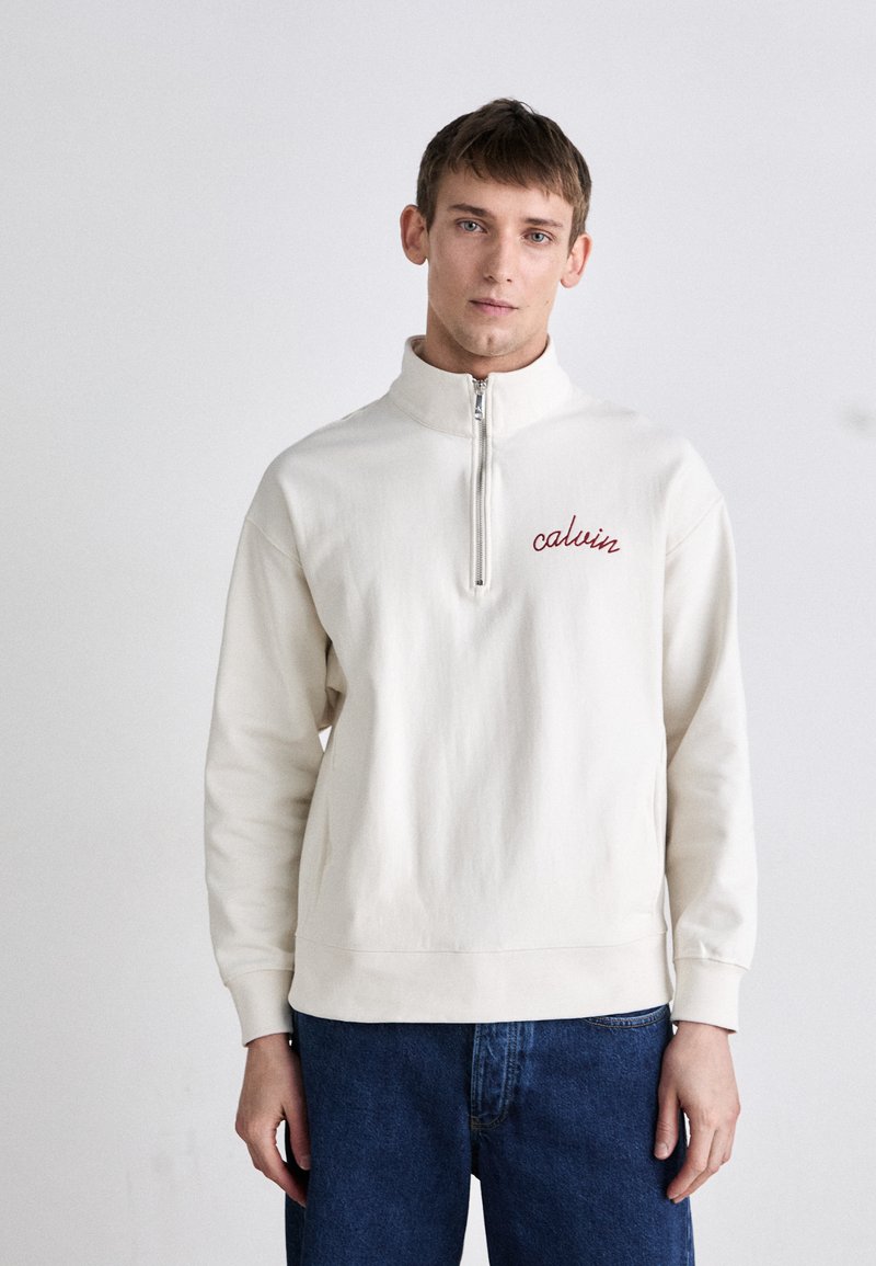 Sudadera de medio cierre color crema con el logotipo bordado en rojo "calvin", fabricada con material suave. Combinada con jeans de mezclilla azul.