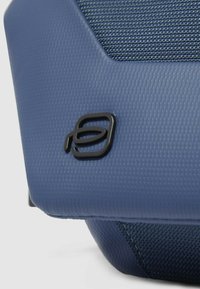 Custodia blu con motivo esagonale e logo nero, che combina materiali tessuti lisci e intrecciati.