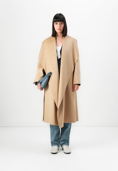 Femeie stând în fața unui fond alb, purtând un palton oversized bej, blugi, pantofi albi, ținând în mâna stângă o geantă albastră.