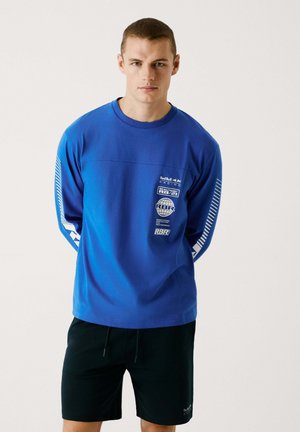 Blue long-sleeve shirt with white graphics and stripes on the sleeves, paired with black shorts. Cotton material with a relaxed fit.