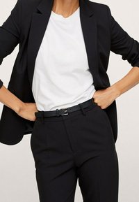 Femme portant un blazer et un pantalon noirs avec un t-shirt blanc rentré, les mains sur les hanches, présentant une tenue professionnelle décontractée.