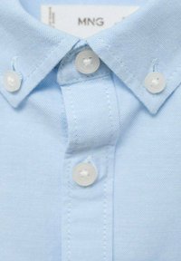 Světle modrá bavlněná košile s límečkem na knoflík, s bílými plastovými knoflíky a jemnou texturou. Obsahuje viditelné detaily šití.