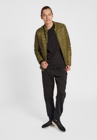 Veste matelassée vert olive avec col montant, portée sur un sweat-shirt noir et un pantalon noir, assortie à des baskets texturées noires.