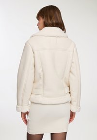 Giubbotto in shearling color crema con ampio colletto, tessuto testurizzato e maniche a polsino. Il giubbotto ha una forma attillata con cuciture laterali.