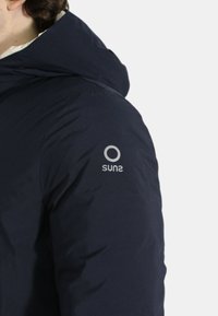 Giacca puffa blu navy con cappuccio, dal tessuto morbido e con un logo argentato sulla manica sinistra. Il design è lineare e moderno.