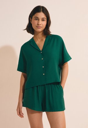 Joven mujer con camisa de manga corta de color verde oscuro con botones y pantalones cortos a juego sobre fondo beige.