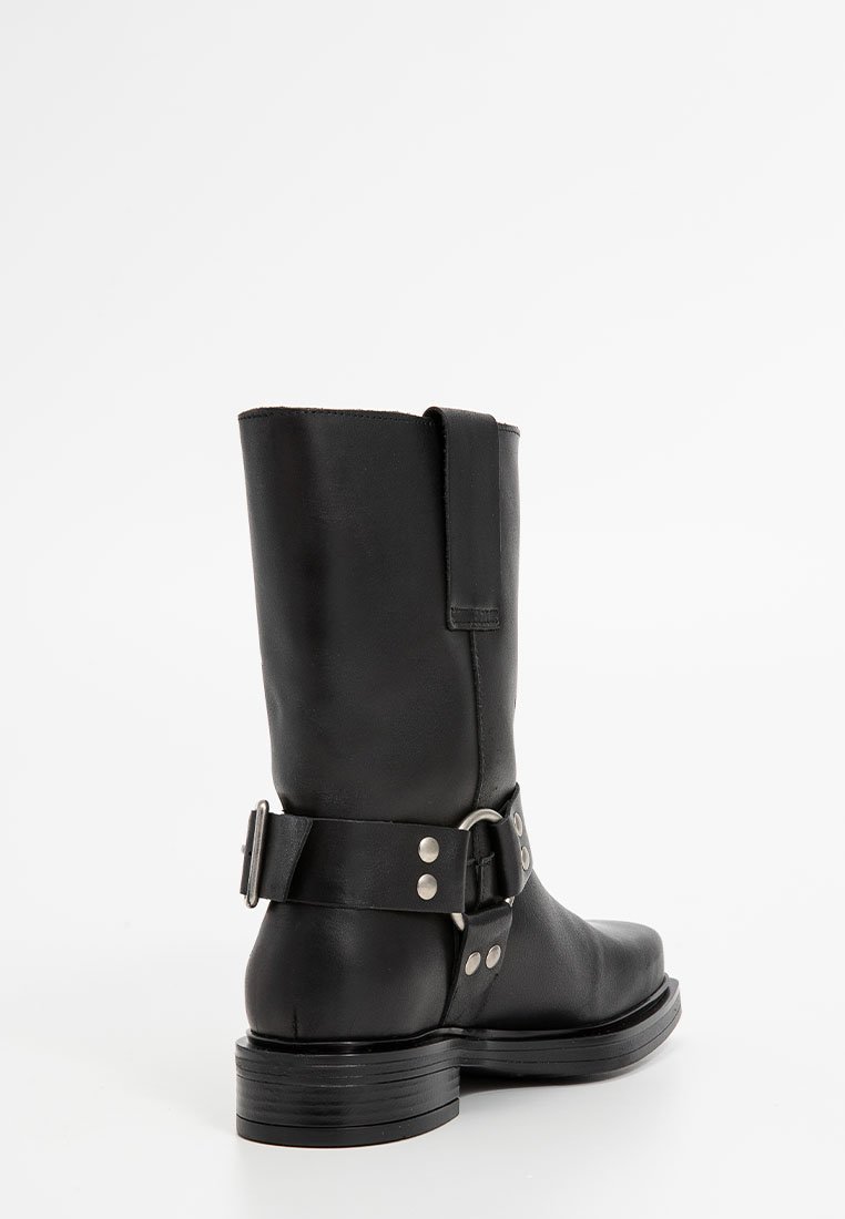 Bottine en cuir noir avec une finition lisse, bout arrondi, boucle en métal argenté et talon empilé, présentant un design simple et épuré.