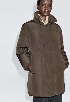 Frau, die einen dunkelbraunen oversized Daunenmantel mit hohem Kragen trägt, die Hände in den Taschen, vor einem schlichten hellen Hintergrund.