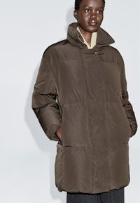 Kvinde iført en mørkebrun oversized puffjakke med høj krave, hænderne i lommerne, mod en ensfarvet lys baggrund.