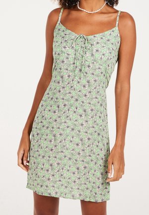 Blomstret mini-kjole i grøn, med tynde stropper, en bindedetalje foran og et fint sort blomsterprint på en lys grøn baggrund.