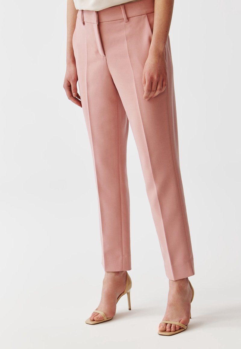 Personne portant un pantalon rose taille haute, coupe droite, et des sandales beiges à talons ouverts, debout devant un fond uni.