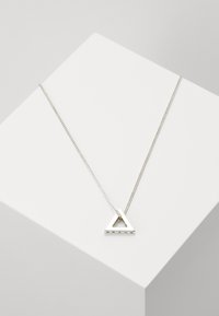 Collier en métal argenté avec un pendentif triangulaire présentant un centre ouvert et un texte gravé. La chaîne est délicate et finement enchaînée.