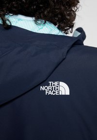 Personne aux cheveux bouclés portant une veste bleu marine montrant le logo The North Face dans le dos, près de la zone de l'épaule.