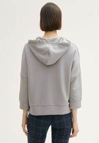 Svijetlosiva hoodie s glatkom teksturom, s nisko postavljenim ramenima i skupljenim kapuljačom. U paru s tamnim hlačama s uzorkom koje imaju dizajn rešetke.