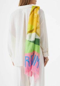 Écharpe colorée drapée sur une chemise blanche texturée. Comprend des teintes jaunes, vertes et roses avec un design graphique et un texte bleu. Les bords sont effilochés.