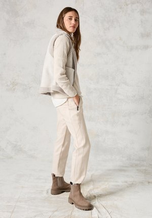 Lichtgrijs fleece vest met een schapenvacht rand, gecombineerd met beige joggingbroek en bruine enkellaarsjes op een gestructureerde vloer.