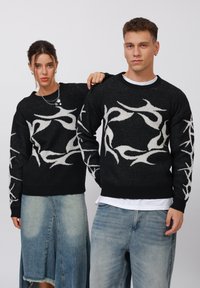 Maglione nero con design astratto bianco, lavorazione a maglia testurizzata, scollatura a girocollo, polsini a costine, indossato con gonne in denim azzurro chiaro e jeans.