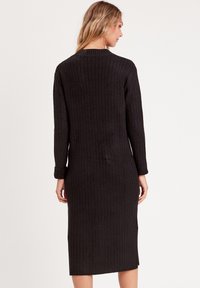 Vestido de punto acanalado negro, de media longitud con manga larga y cuello redondo. Presenta un diseño ajustado y aberturas laterales para mayor libertad de movimiento.