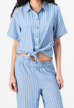 Frau trägt ein hellblau-weiß gestreiftes, kurzärmliges Hemd mit Knöpfen und vertikalen Streifen, das an der Taille gebunden ist, sowie passende Hosen mit elastischem Bund.