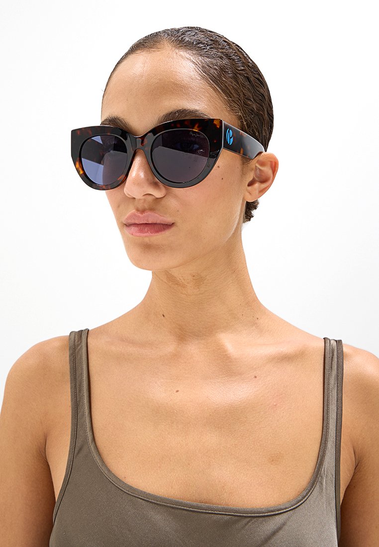 Frau mit zurückgegeltem Haar, die eine Schildpatt-Katze-Augen-Sonnenbrille und ein braunes ärmelloses Oberteil vor einem weißen Hintergrund trägt.
