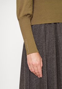 Olijfgroene geribbelde trui met lange mouwen, voorzien van metalen knoopaccenten bij de manchetten, gecombineerd met een getextureerde donkere rok.