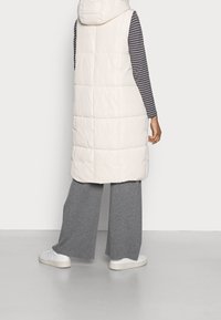 Vadderad gräddfärgad väst med huva, med kviltade paneler. Kombinerad med grå byxor med vida ben och vita sneakers. Randsydd långärmad tröja under.