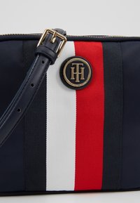 Laosikangas sininen laukku, jossa on kultainen logo ja punavalkoinen keskiraita. Säädettävä hihna on tummansinistä nahkaa ja siinä on metallinen solki.