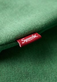 Groene stof met een zachte textuur, voorzien van een rood label met "Superdry" in witte cursieve tekst. Close-up van de details van het materiaal en het label.