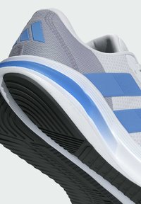Chaussure de sport blanche avec une tige en mesh gris, des accents bleus et une semelle extérieure en caoutchouc noir texturé. Présente trois rayures bleues sur le côté.