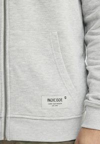 Sivi zip-up dukserica s teksturiranim materijalom, rebrastim manžetama i prednjim džepom. Etiketa glasi "INDICODE CARRY OUR PASSION EST. 1993."