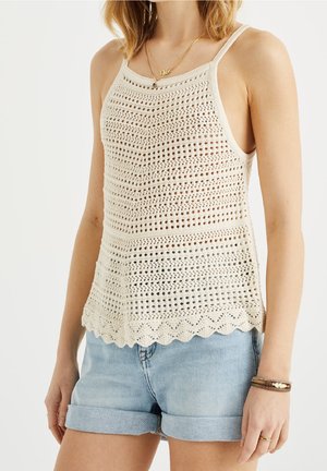 Kobieta ubrana w kremowy szydełkowany bezrękawnik z falistym brzegiem i jasnoniebieskie podwinięte szorty dżinsowe na neutralnym tle.