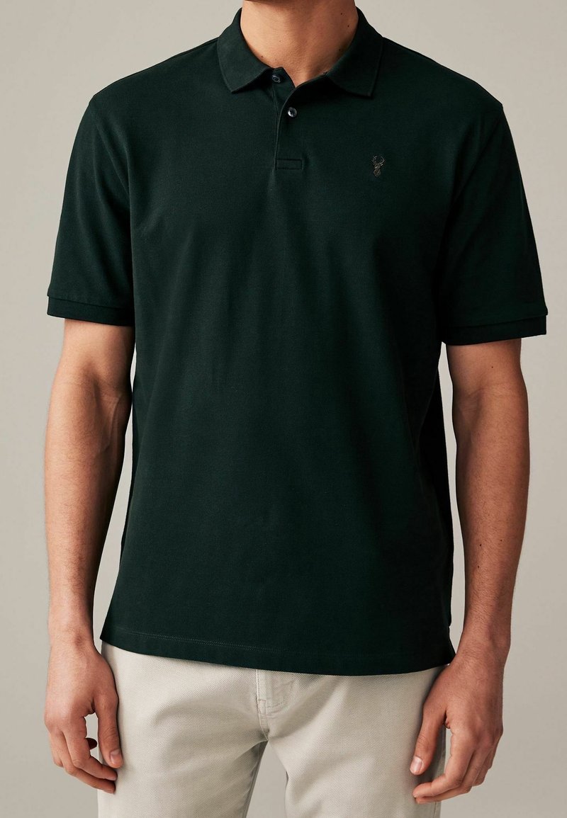 Polo noir manches courtes avec col et deux boutons, porté avec un pantalon clair, montrant le haut du corps et les bras.