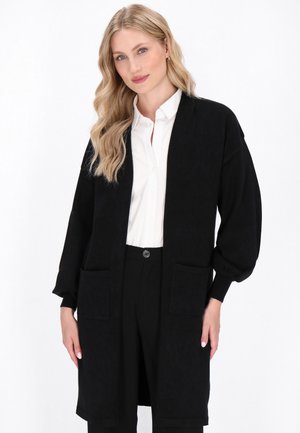 Schwarzer langer Cardigan aus weichem Stoff, mit lockerer Passform, langen Ärmeln, zwei vorderen Taschen und gerippten Bündchen. Über einem weißen Hemd gestylt.