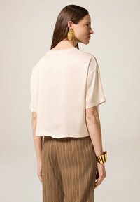 T-shirt corto di un beige chiaro con maniche corte, texture liscia e scollatura rotonda, abbinato a pantaloni marroni a righe e orecchini dorati.