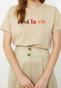 Beige katoenen T-shirt met korte mouwen en "c'est la vie" bedrukt in bruin, rood en oranje. Gecombineerd met hooggetailleerde beige broeken met knoopdetails.