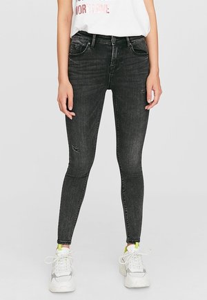 Femme portant un jean skinny noir usé et des baskets blanches épaisses, debout devant un fond clair uni.